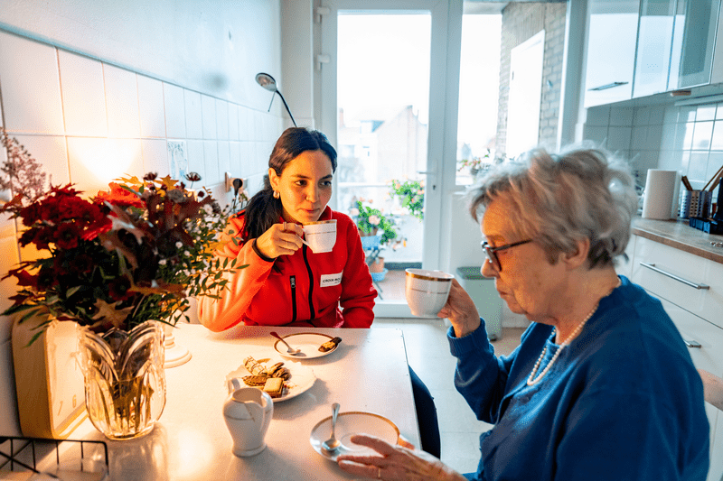 Une bénévole de la CRB et une dame âgée son assises autour d'une table autour d'un café et de petits gâteaux